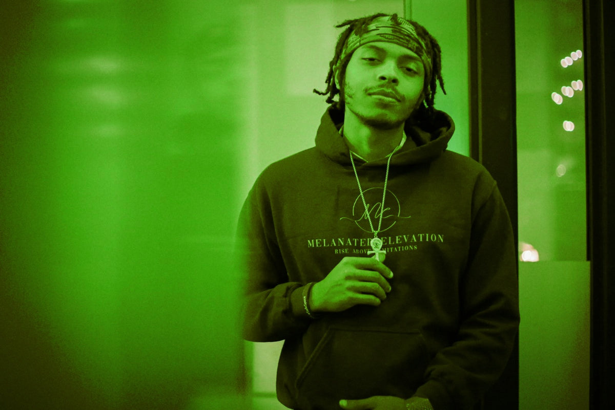 Founder wearing a black Melanated Elevation hoodie in a green-lit editorial portrait