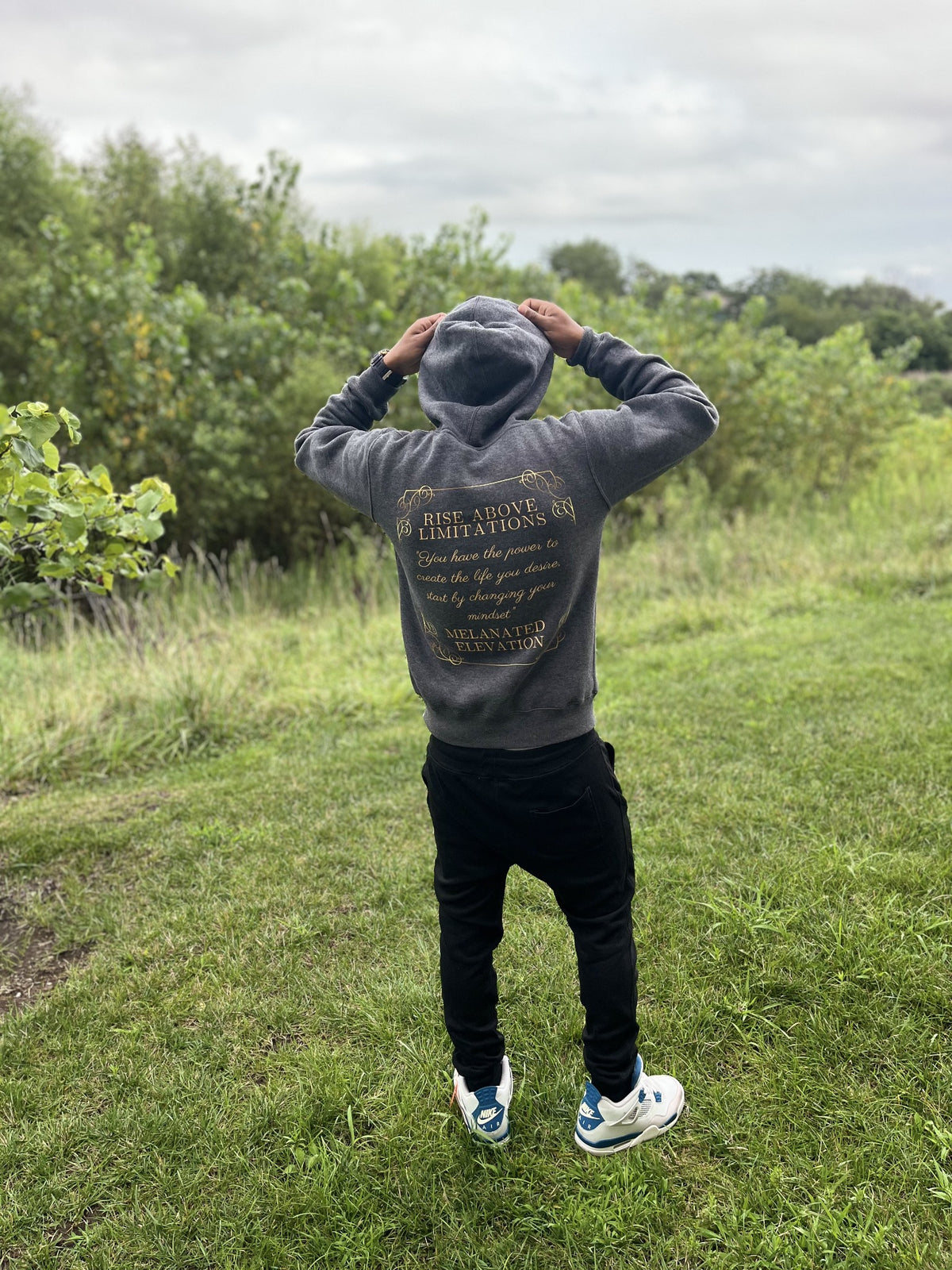 Model outdoors wearing the gray Melanated Elevation hoodie with the back message visible